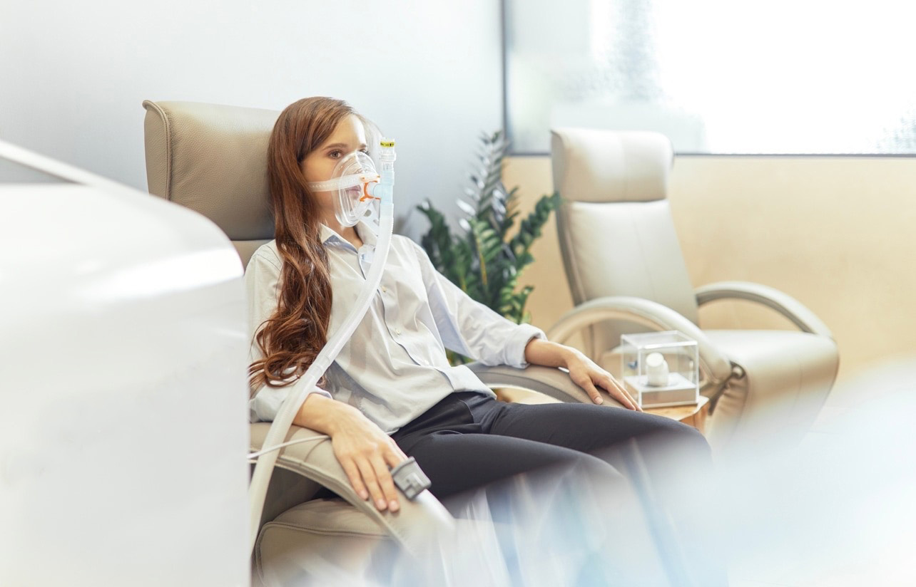 A woman sits back in a reclining chair breathing through a clear soft mask connected by a ribbed hose to a console, a pulse oximeter clipped to her finger, in a bright clinic lounge with a plant and daylight behind her.